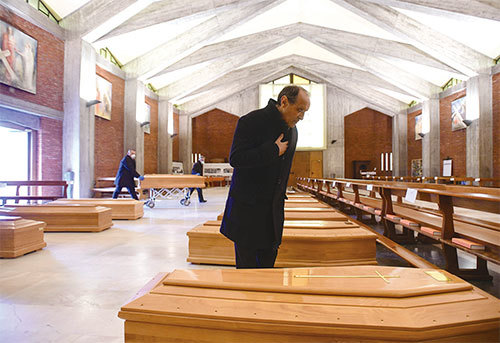 2020年3月26日，意大利處於封城時期，一座教堂裡存放著感染中共病毒死亡者的棺材，等待被軍車運走。神父站在其中一棺材旁禱告。（AFP）